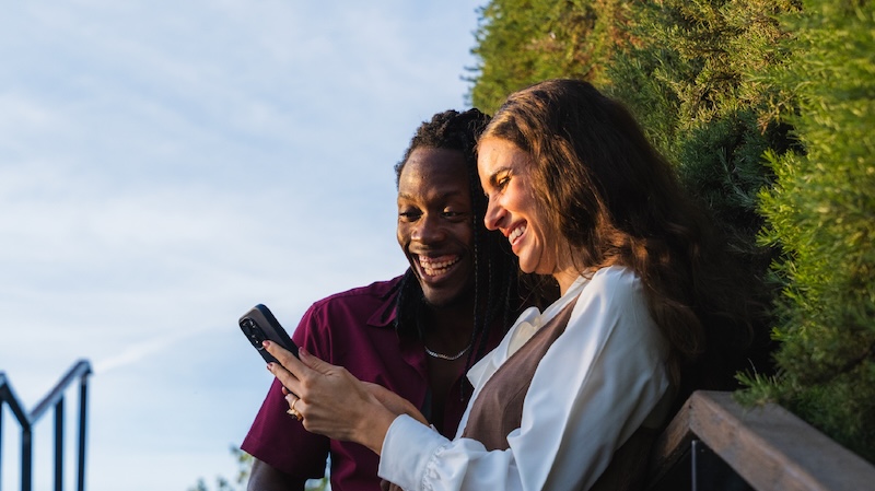 Man and woman smiling while looking at her cellphone, standing outdoors